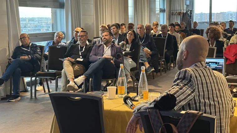 Il pubblico seduto ascolta il relatore, in primo piano le bottigliette e i tavolini